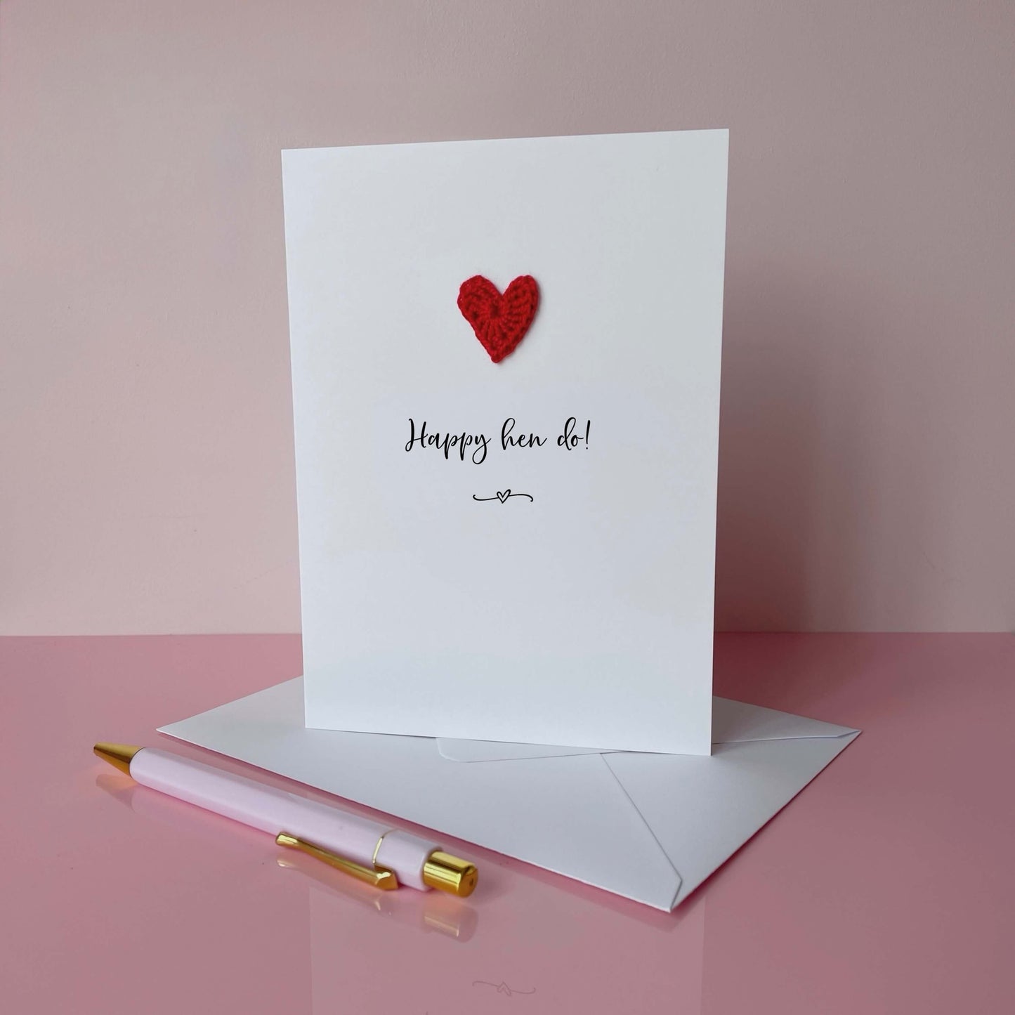 Greeting card with red heart and 'Happy hen do!' text on a pink surface with a pen and envelope.