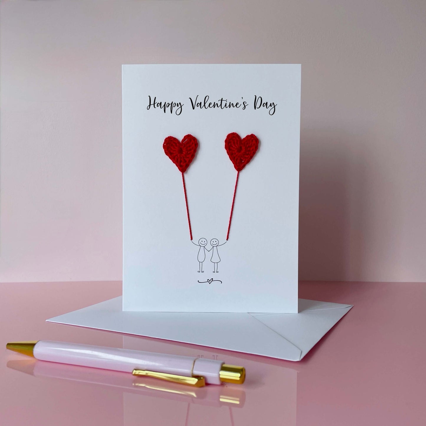 A white card featuring a line drawn couple holding hands and each holding a red crochet heart balloon.  ‘Happy Valentine’s Day’ is printed at the top of the card in black cursive text.