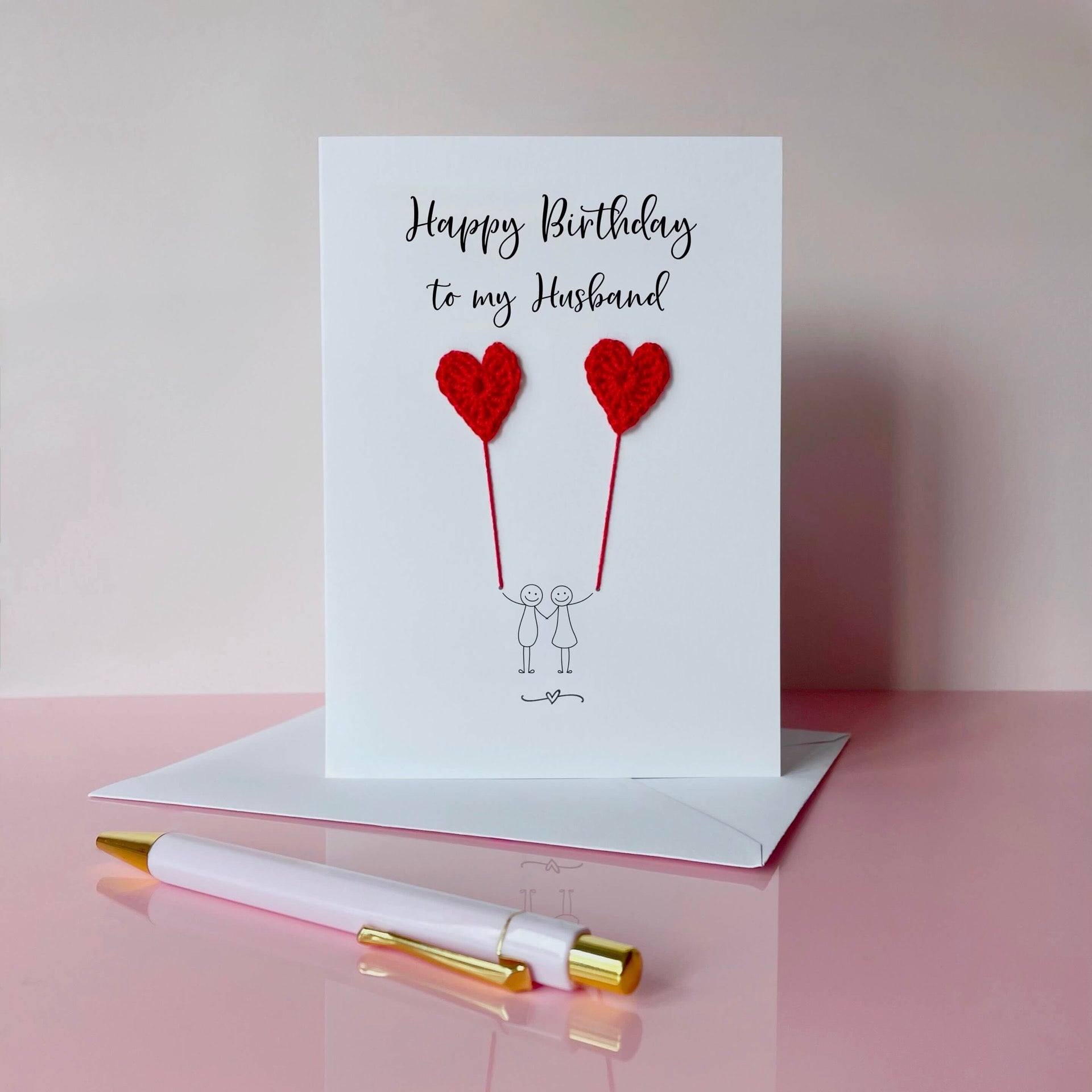 A white birthday card featuring a line drawn couple holding hands and each holding a red crochet heart balloon.  ‘Happy Birthday to my Husband’ is printed at the top of the card in black cursive text.