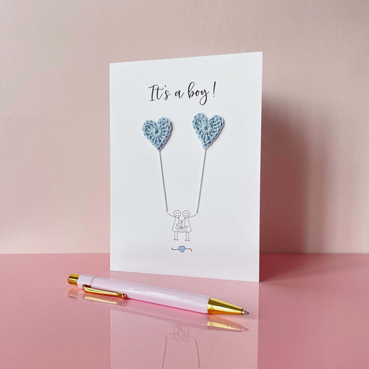 A simple greeting card with pale blue crochet heart balloons and 'It's a boy!' text. A line drawn couple are on the card  - they are holding a baby carrier. 