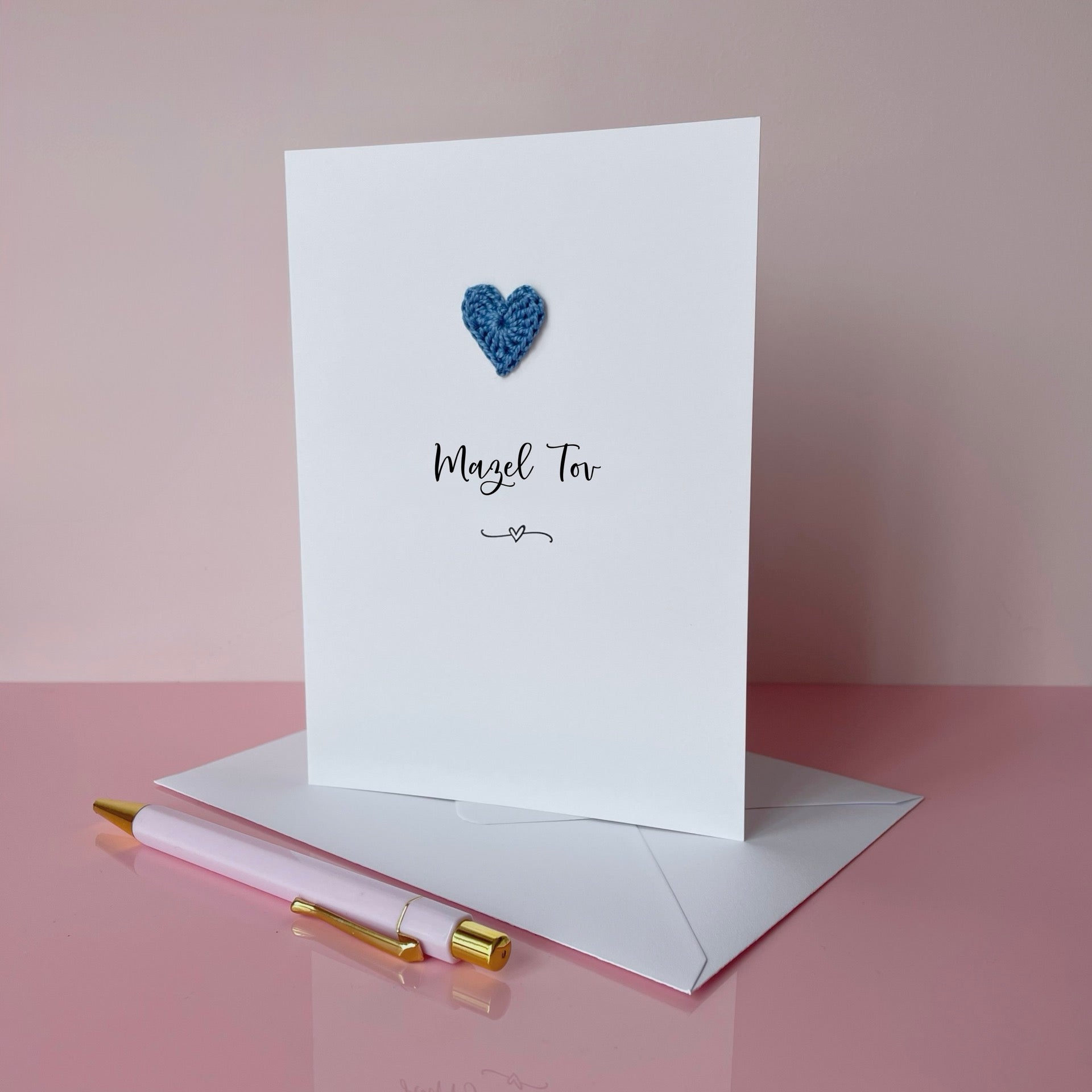 Greeting card with a blue crochet heart and text on a pink background, 'Mazel Tov' is printed on the card in lovely black cursive text. 