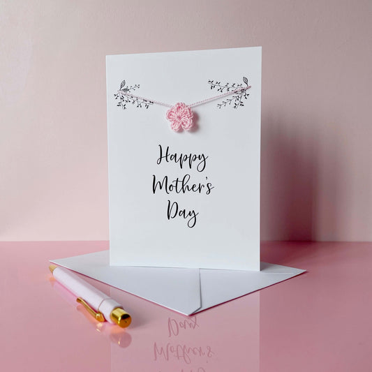 A white card with 'Happy Mother's Day' printed in the centre in black cursive text. A pale pink crochet flower has been strung across the front of the card using pale pink yarn. Printed black foliage appears at the top of the card. 