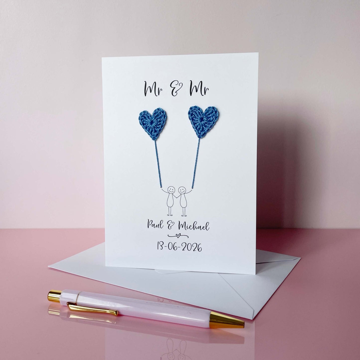 A white card featuring a line drawn couple holding hands and each holding a blue crochet heart balloon. ‘Mr & Mr’ is printed at the top of the card and the couples names and wedding date is printed just below the couple. 