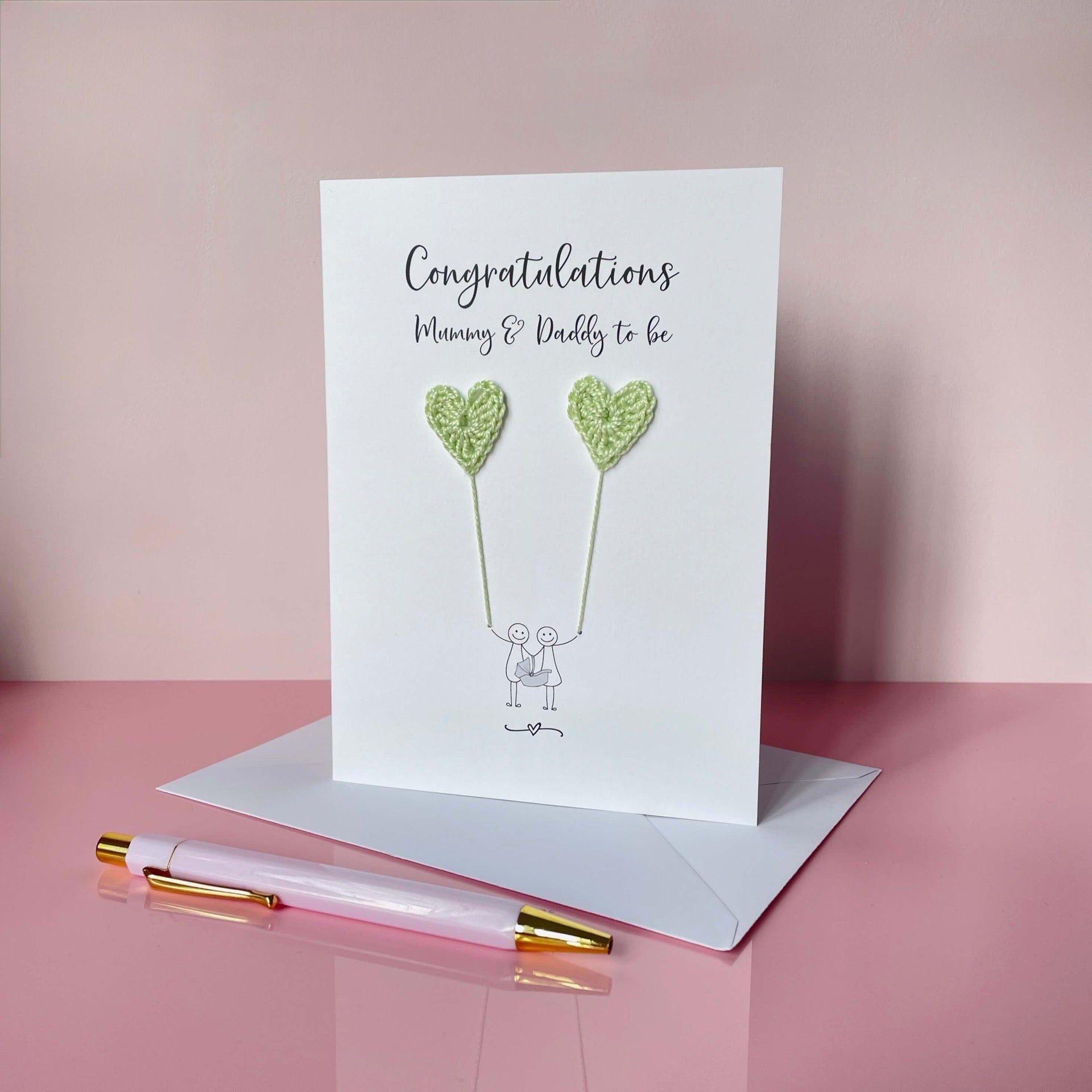 A white card with 'Congratulations Mummy & Daddy to be' printed at the top of the card. An illustrated couple holding a baby carrier are each holding a pale green crochet heart balloon. 