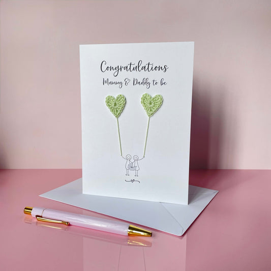 A white card with 'Congratulations Mummy & Daddy to be' printed at the top of the card. An illustrated couple holding a baby carrier are each holding a pale green crochet heart balloon. 