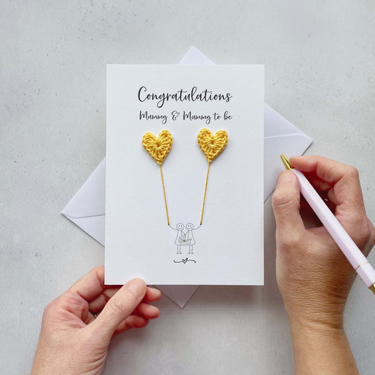 A white card with 'Congratulations Mummy & mummy to be' printed at the top. the illustrated couple are each holding a yellow crochet heart, this looks like they are holding balloons. 