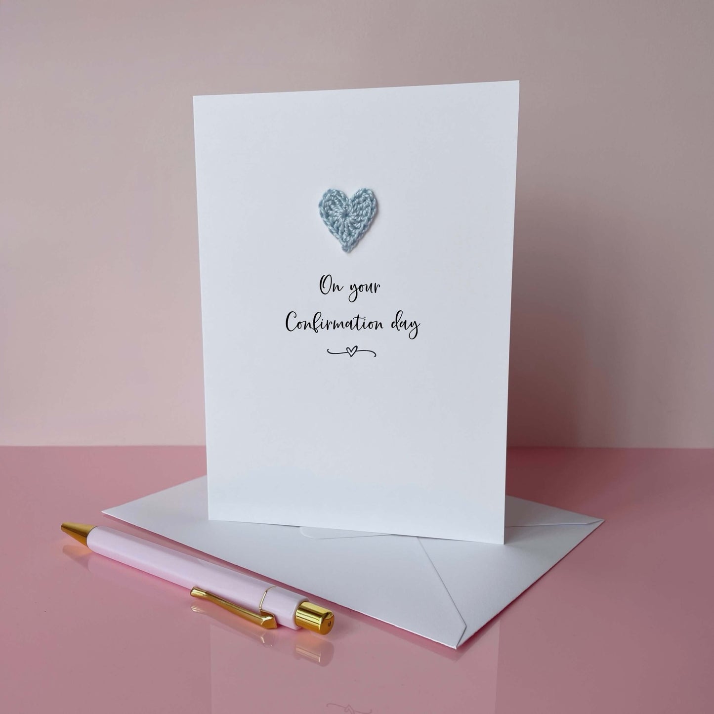 Greeting card with a pale blue crochet heart and text on a pink background, 'On your confirmation day' is printed on the card in lovely black cursive text. 