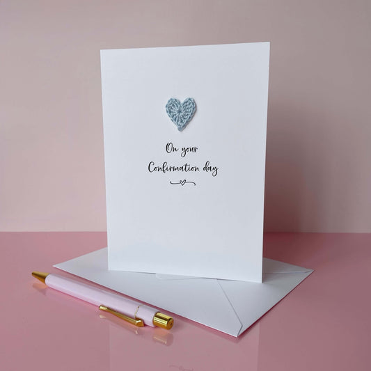 Greeting card with a pale blue crochet heart and text on a pink background, 'On your confirmation day' is printed on the card in lovely black cursive text. 