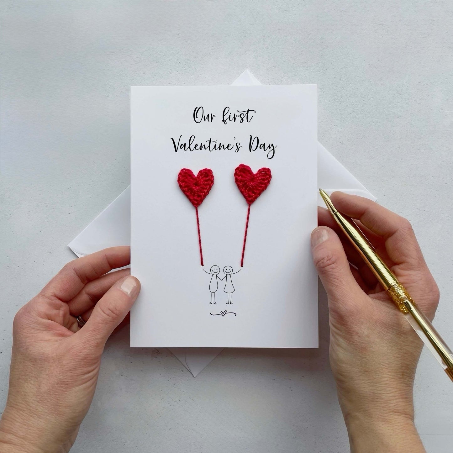 Card with red crochet hearts and text 'Our first Valentine's Day' held by hands on a light gray background.