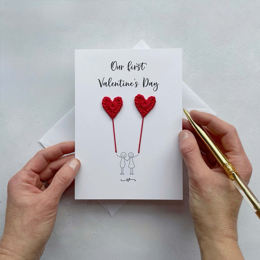 Card with red crochet hearts and text 'Our first Valentine's Day' held by hands on a light gray background.