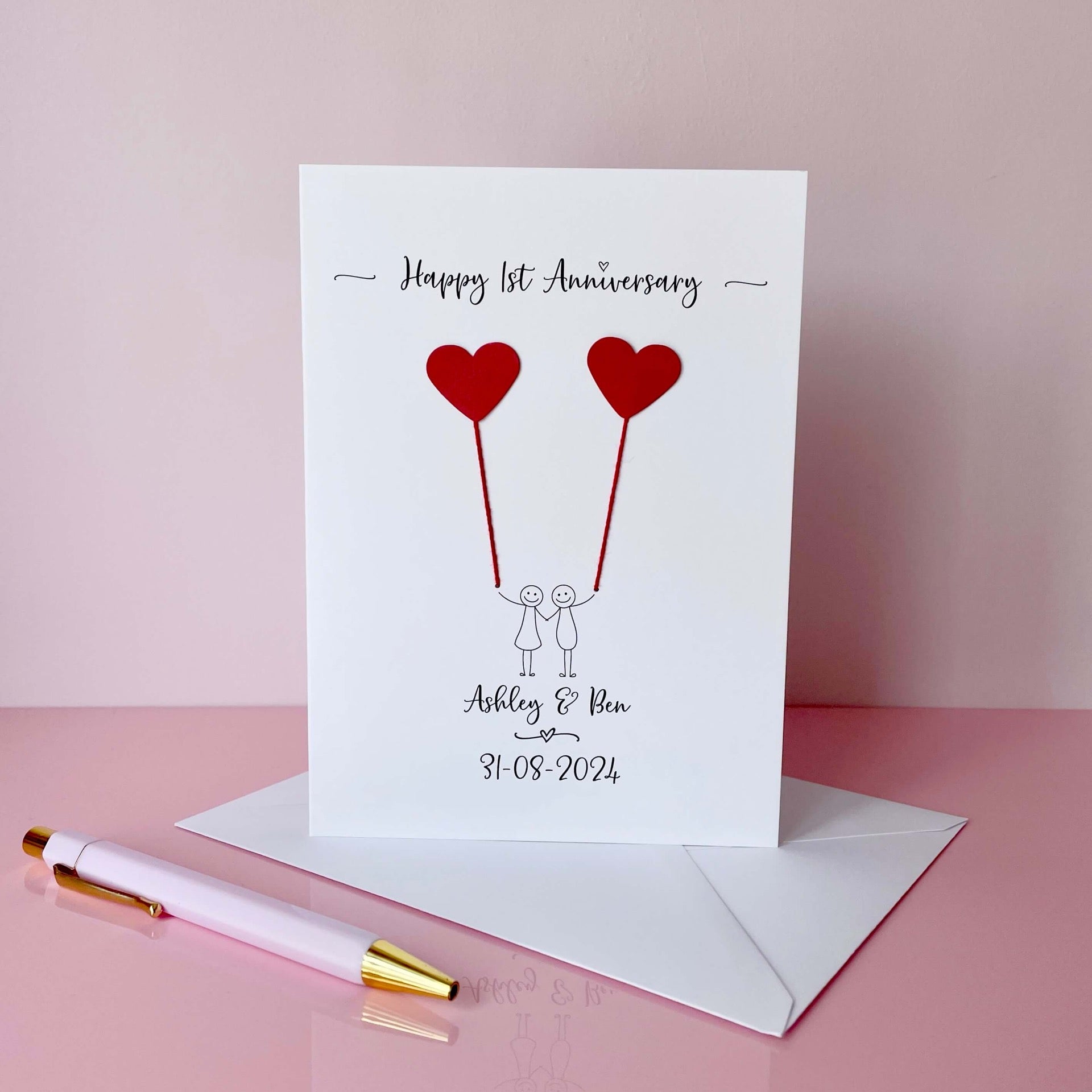 A white card featuring a line drawn couple each holding a red heart made from cardboard. Happy 1st Anniversary is printed at the top of the card and the couples names and wedding date are printed at the bottom. 