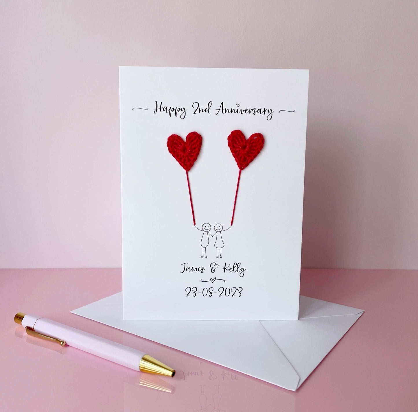 A white card featuring a line drawn couple each holding a red crochet heart made from cotton yarn. Happy 2nd Anniversary is printed at the top of the card and the couples names and wedding date are printed at the bottom. 
