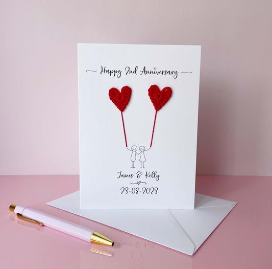 A white card featuring a line drawn couple each holding a red crochet heart made from cotton yarn. Happy 2nd Anniversary is printed at the top of the card and the couples names and wedding date are printed at the bottom. 
