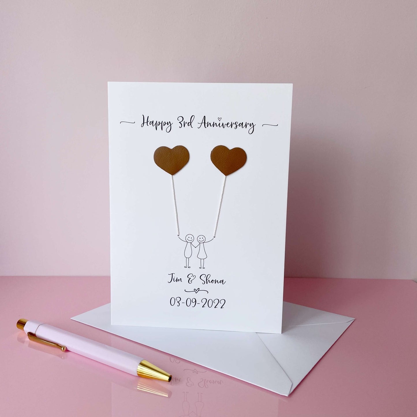 A white card featuring a line drawn couple each holding a leather heart shaped balloon. Happy 3rd Anniversary is printed at the top of the card and the couples names and wedding date are printed at the bottom. 