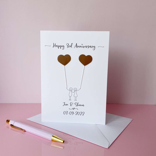 A white card featuring a line drawn couple each holding a leather heart shaped balloon. Happy 3rd Anniversary is printed at the top of the card and the couples names and wedding date are printed at the bottom. 