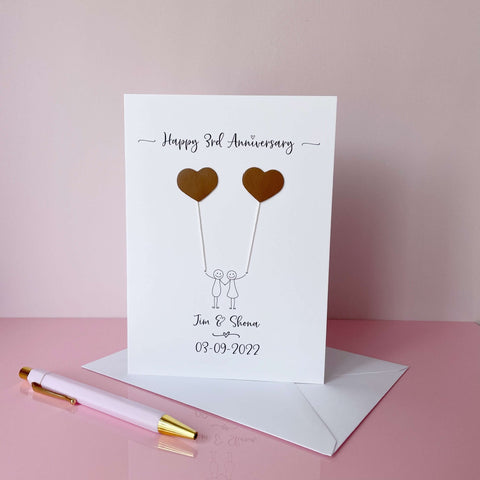A white card featuring a line drawn couple each holding a leather heart shaped balloon. Happy 3rd Anniversary is printed at the top of the card and the couples names and wedding date are printed at the bottom. 