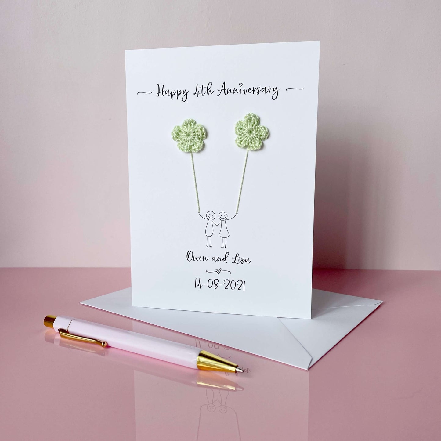 4th Anniversary card with floral crochet hearts and personalised text on a pink surface with a pen and envelope.