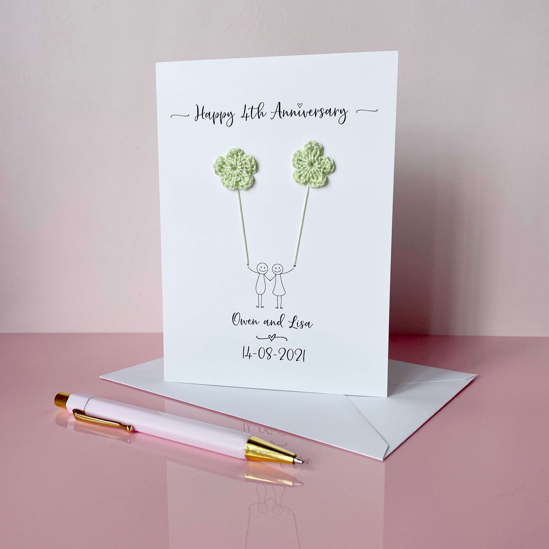 4th Anniversary card with floral crochet hearts and personalised text on a pink surface with a pen and envelope.