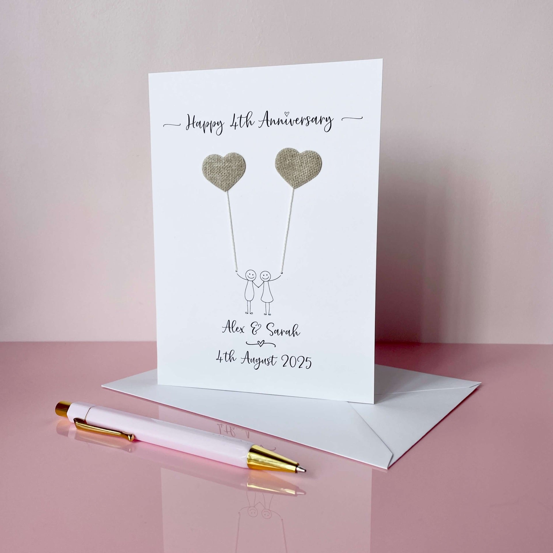 A white card featuring a line drawn couple each holding a heart shaped balloon which is made from linen. Happy 4th Anniversary is printed at the top of the card and the couples names and wedding date are printed at the bottom. 