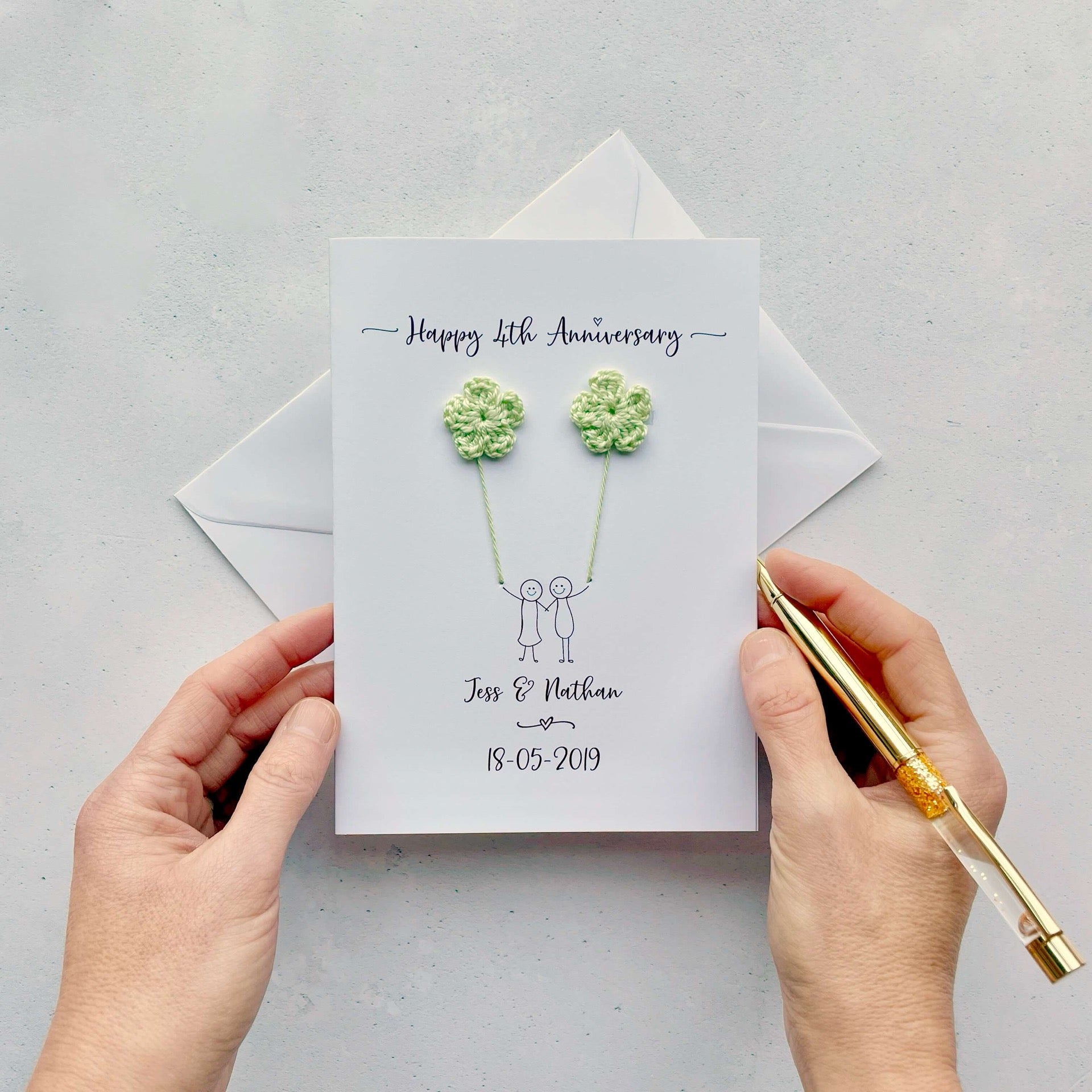 Handmade 4th flower anniversary card embellished with 2 mini cotton crochet flowers, both attached by cotton yarn, resembling balloons being held by a couple underneath. Happy 4th Anniversary is printed at the top, the couples names/ wedding date at the bottom.