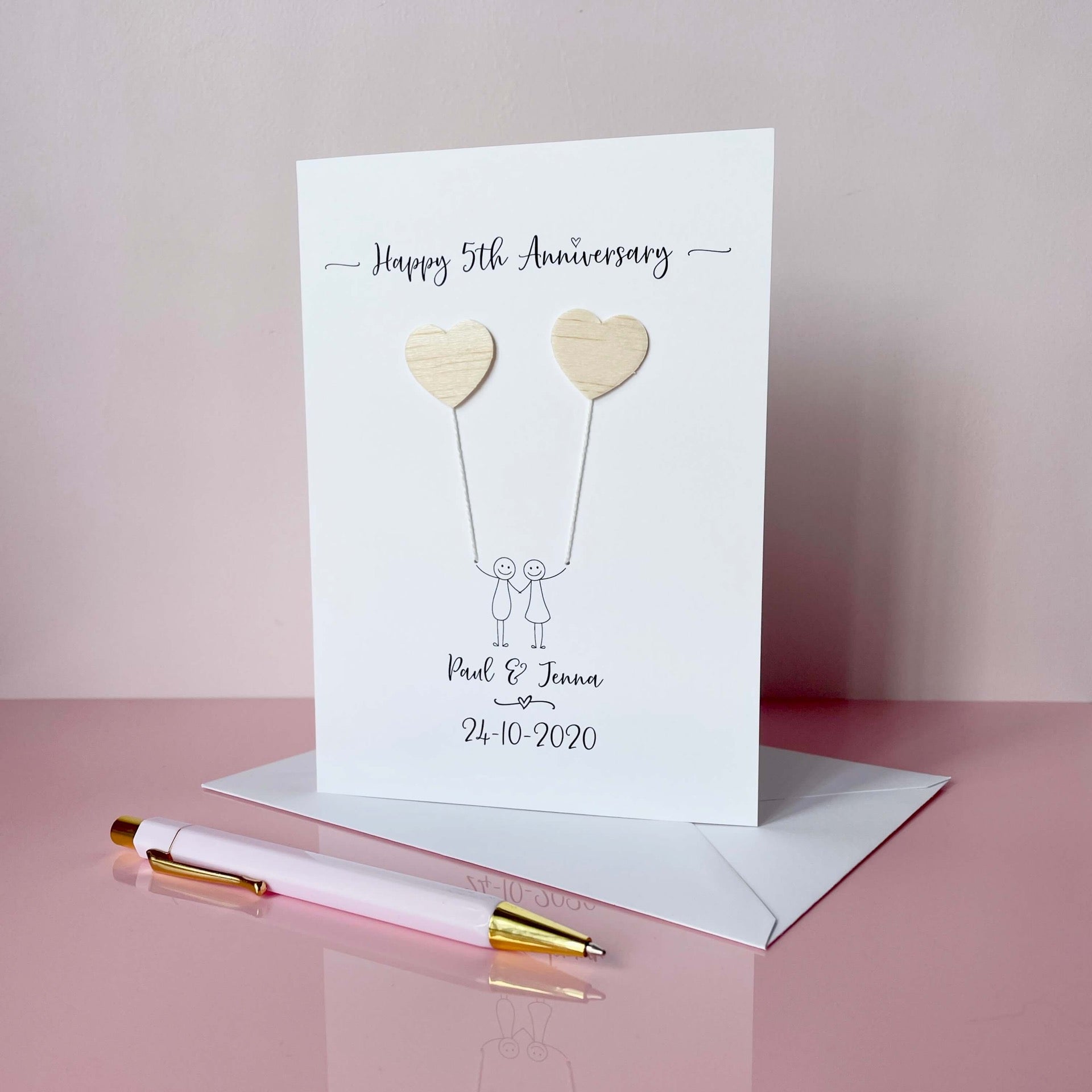 A white card featuring a line drawn couple holding hands and each holding a heart shaped balloon which is made from balsa wood. Happy 5th Anniversary is printed at the top of the card and the couples names and wedding date are printed at the bottom.