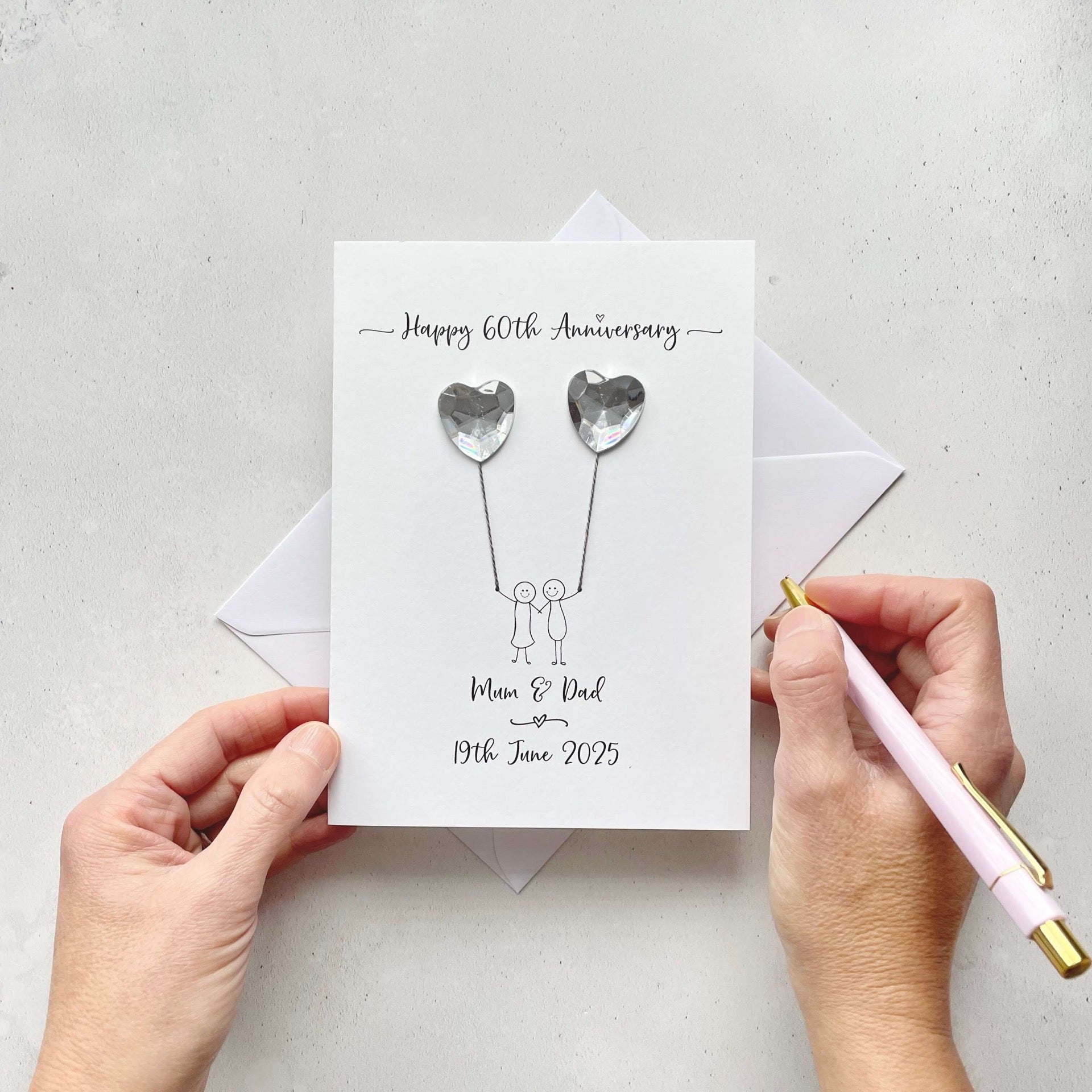 Handmade Diamond anniversary card embelished with 2 heart shaped plastic gems, both attached by cotton yarn, resembling balloons being held by a couple underneath. Happy 60th Anniversary is printed at the top, the couples names/ wedding date at the bottom.