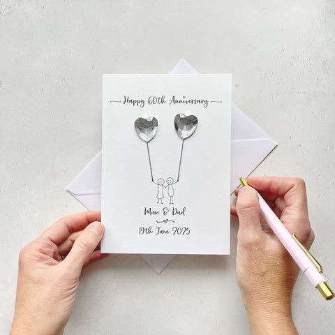 Handmade Diamond anniversary card embelished with 2 heart shaped plastic gems, both attached by cotton yarn, resembling balloons being held by a couple underneath. Happy 60th Anniversary is printed at the top, the couples names/ wedding date at the bottom.