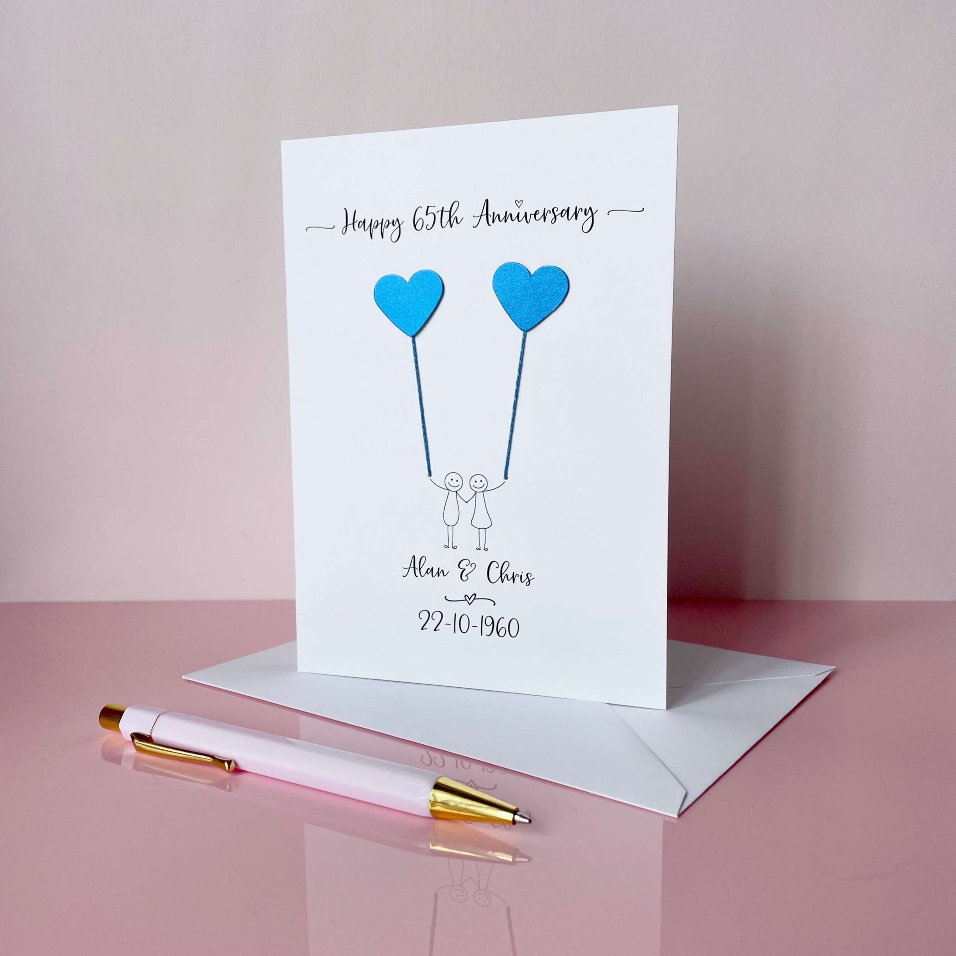 A white card featuring a simple line drawn couple each holding a blue pearlised heart shaped card balloon. Happy 65th Anniversary is printed at the top and the couples names and wedding date is printed at the bottom of the card. 