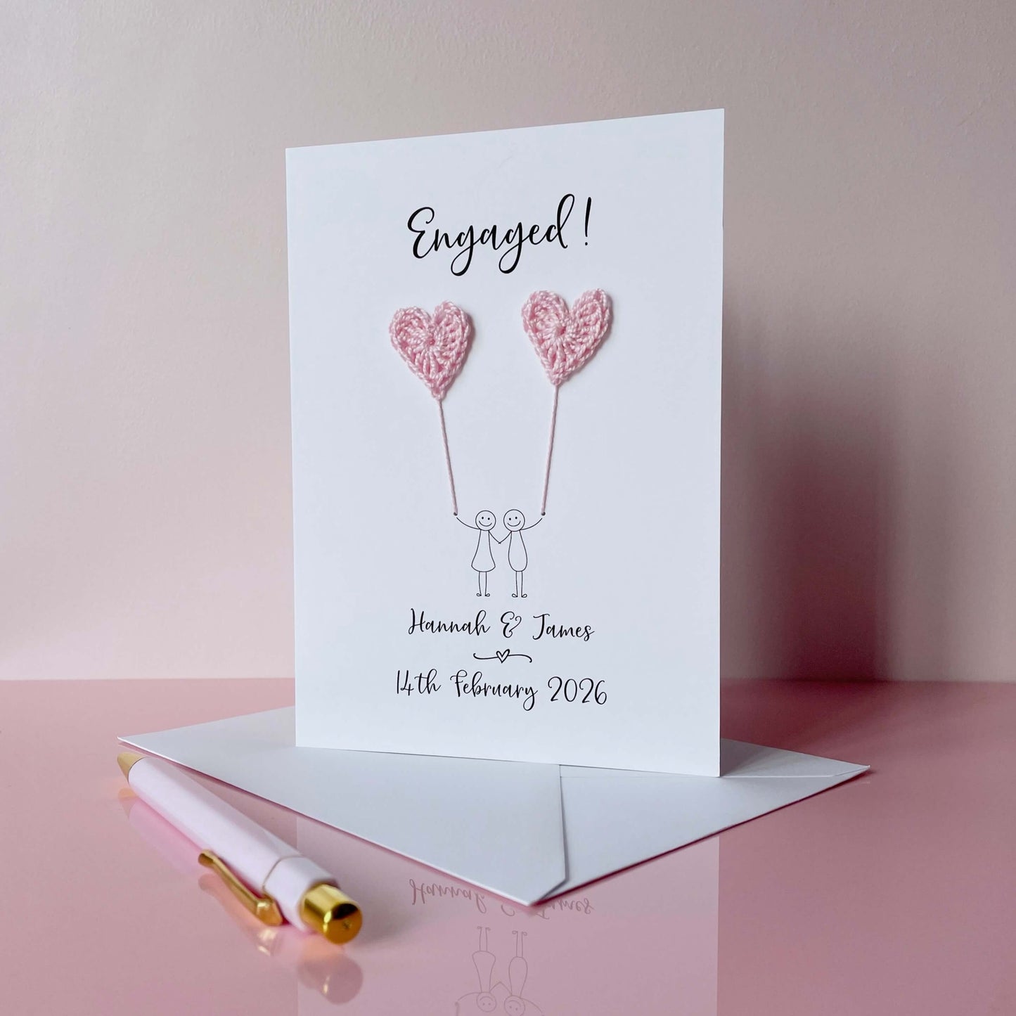 A personalised engagement card with two pale pink crochet hearts. Engaged! is printed at the top of the card in black cursive text and the couples names and engagement date are printed at the bottom. 