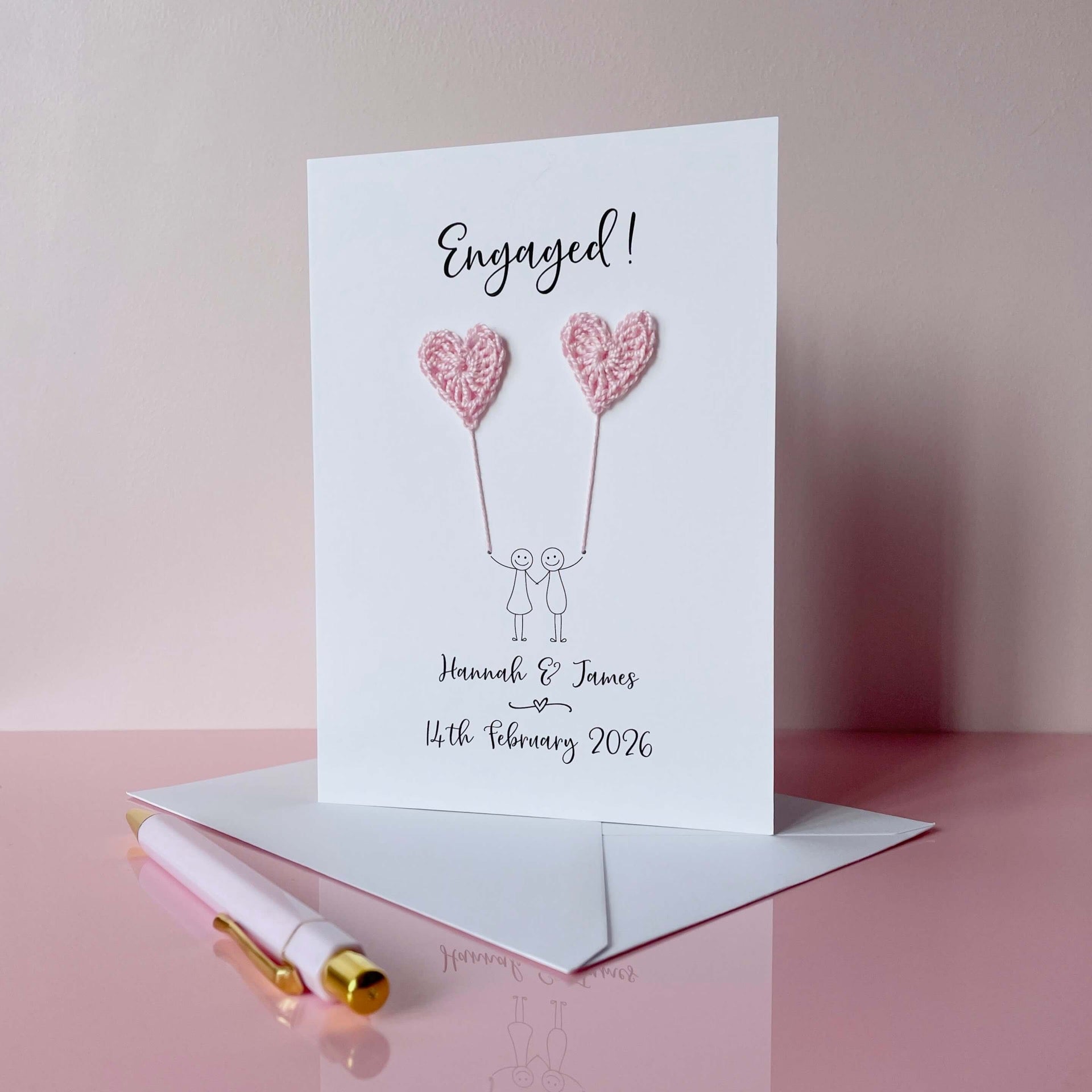 A personalised engagement card with two pale pink crochet hearts. Engaged! is printed at the top of the card in black cursive text and the couples names and engagement date are printed at the bottom. 