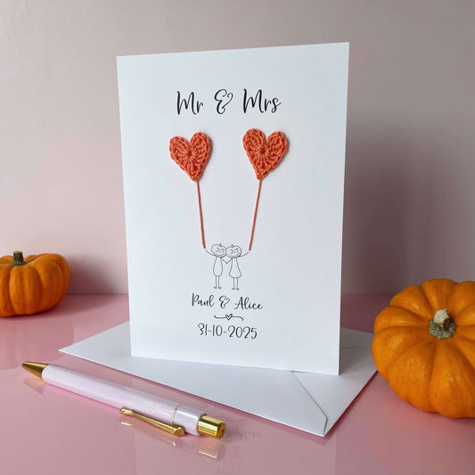 A white card featuring a line drawn couple holding hands and each holding an orange (coral) crochet heart balloon. ‘Mr & Mrs’ is printed at the top of the card and the couples names and wedding date is printed just below the couple. The couple have pumkins as heads and heart shaped cut out eyes. 