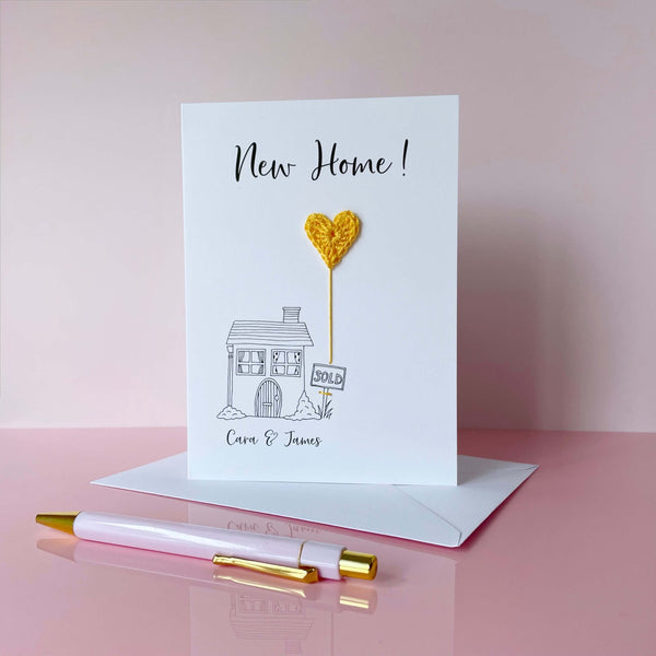 A white card with line drawn house on the front. A yellow crochet heart has been attatched to the card to look like  a heart shaped balloon coming up from the chimney. ’New Home!’ is printed at the top of the card in black cursive text. The couples names have been printed below the house.  