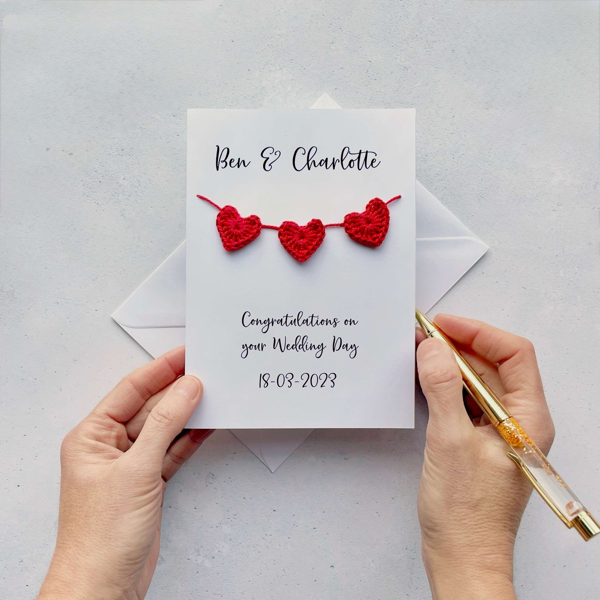 A white card with a string of 3 red crochet hearts across the front. Above the hearts are the two names and at the bottom of the card is 'Congratulations on your wedding day' and the wedding date. 