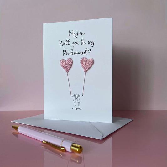Greeting card with crochet heart balloons and text 'Megan, Will you be my Bridesmaid?' on a pink background.