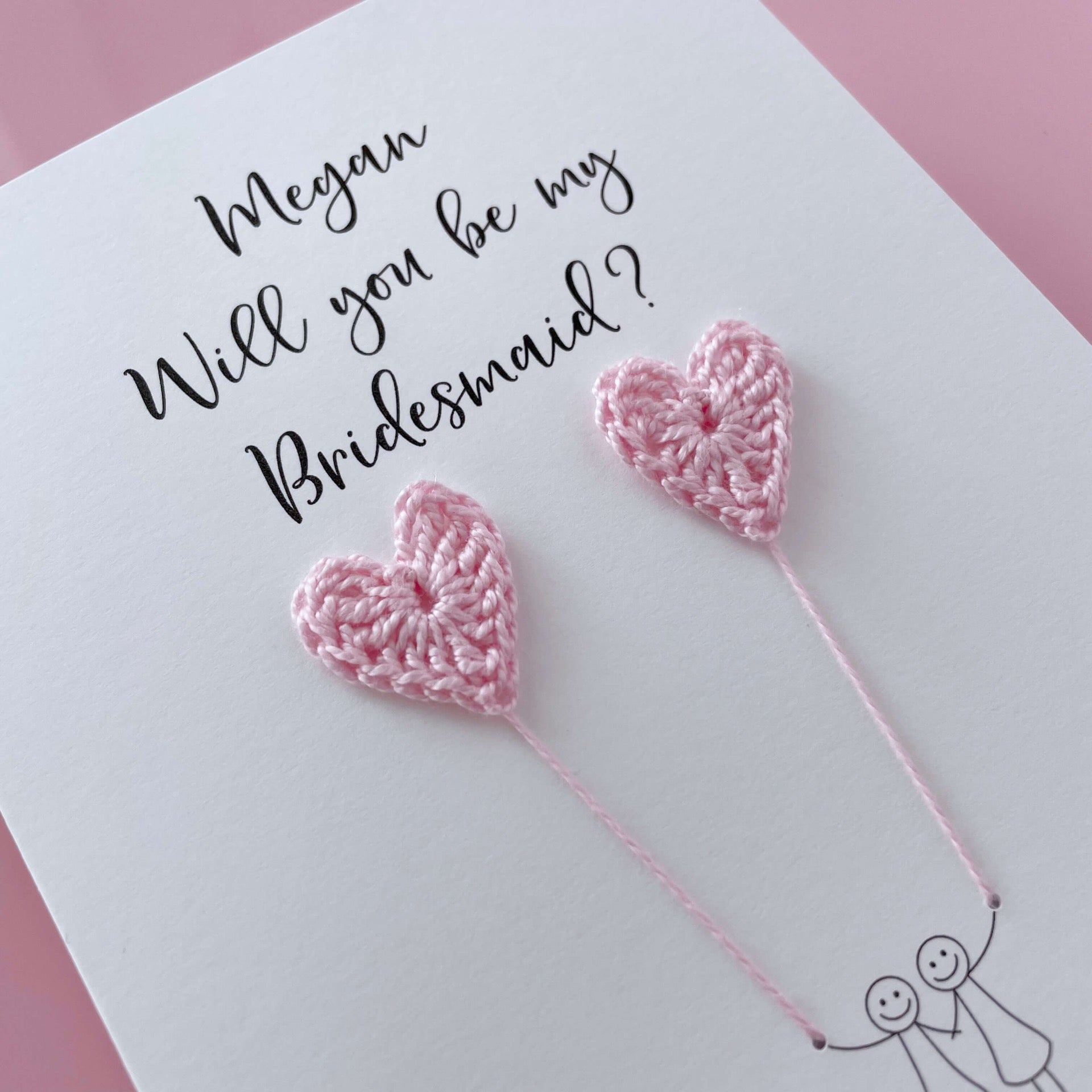 A white card with a line drawn couple printed on it - the couple are each holding a pale pink crochet heart balloon. Will you be my Bridesmaid is printed at the top of the card with the option of adding a name, making it the perfect keepsake. 