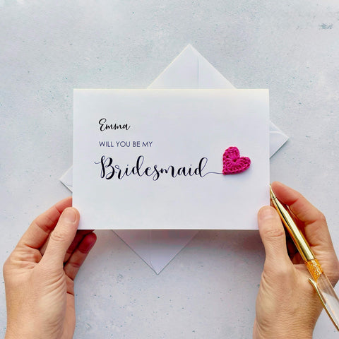 A white card with ‘Will you be my Bridesmaid’ printed on it. The word ‘Bridesmaid’ appears larger than the rest of the text and it has a little bright pink crochet heart at the end of the word.  The name ‘Emma’ has been printed at the top of the text, making this card a lovely keepsake. 