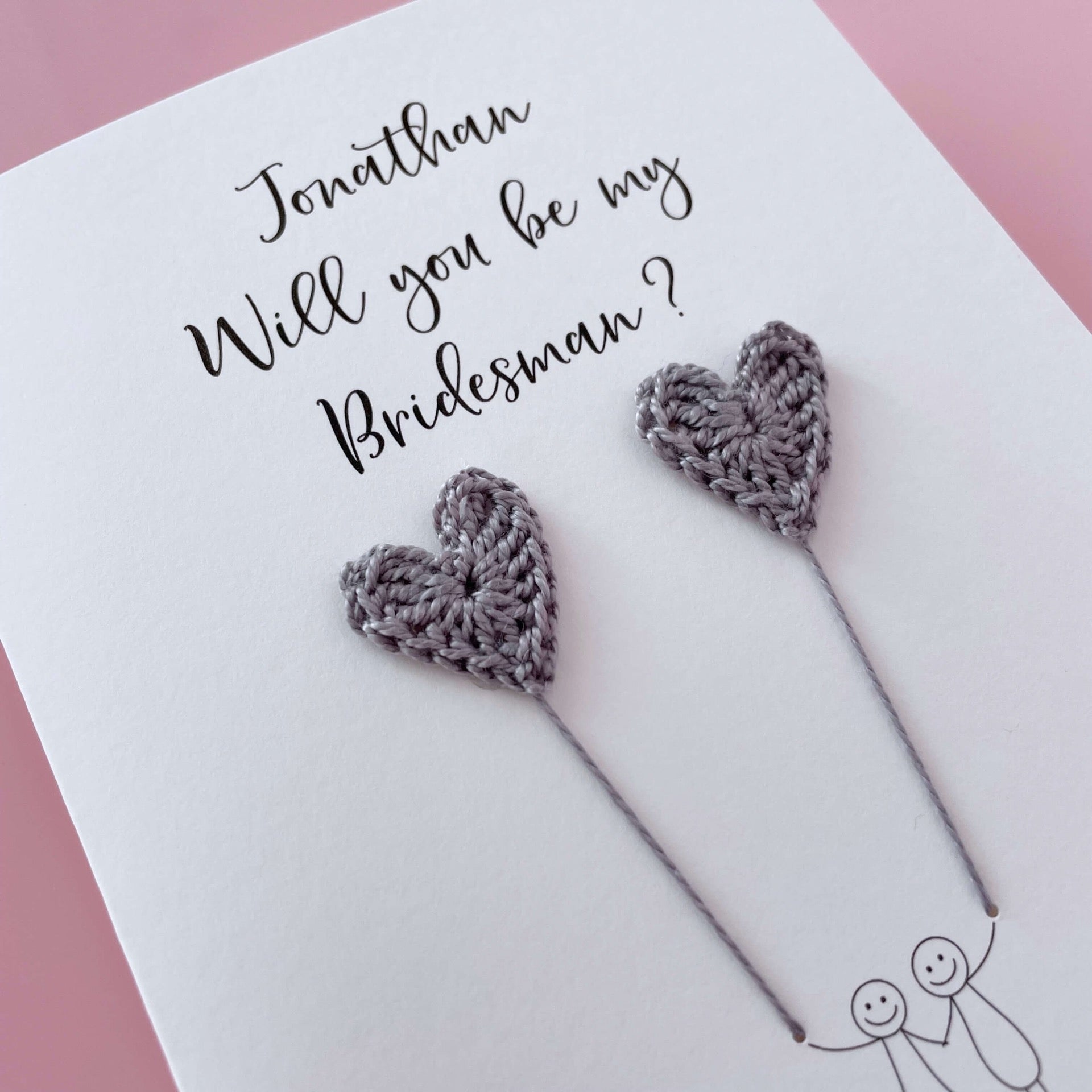 A white card with a line drawn couple printed on it - the couple are each holding a grey crochet heart balloon. Will you be my Bridesman is printed at the top of the card with the option of adding a name, making it the perfect keepsake. 