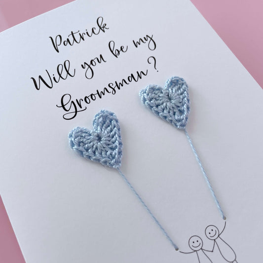 A white card with a line drawn couple printed on it - the couple are each holding a pale blue crochet heart balloon. Will you be my Groomsman is printed at the top of the card with the option of adding a name, making it the perfect keepsake. 