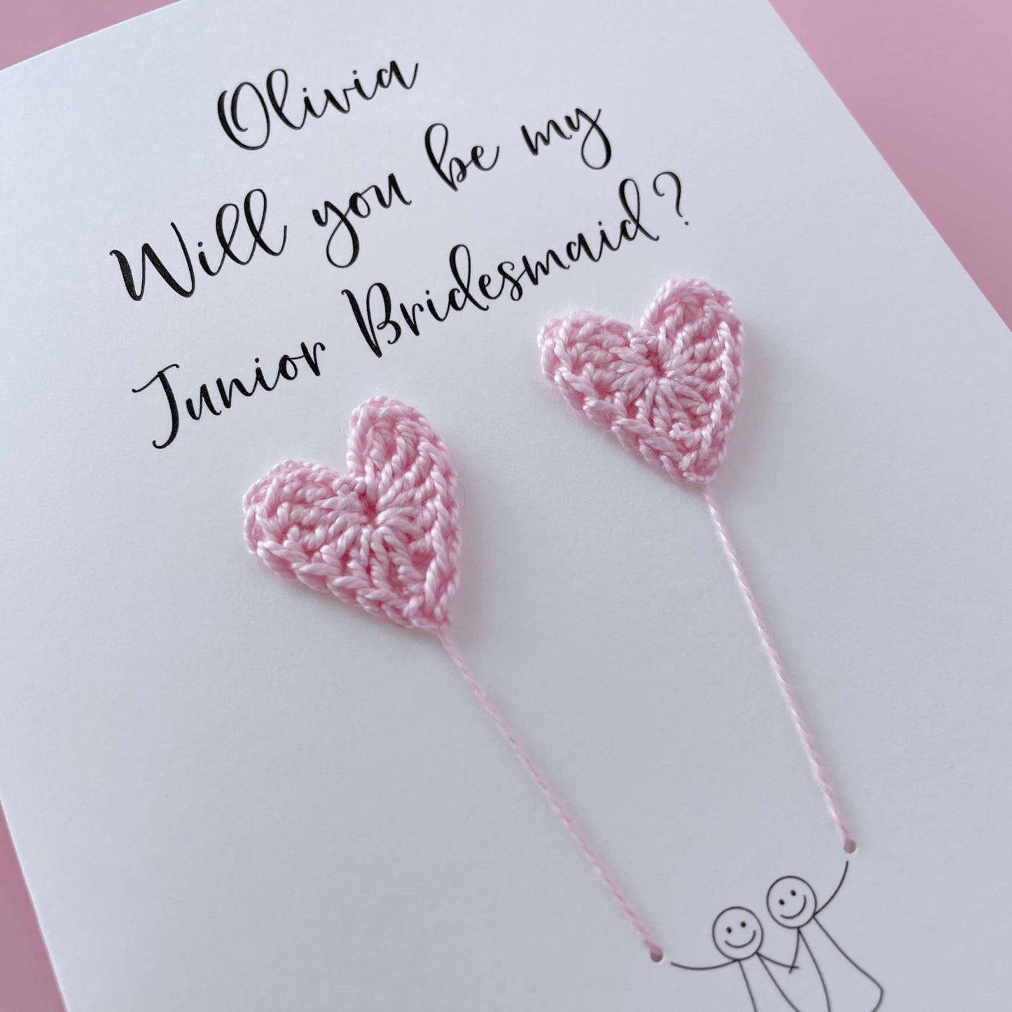 A white card with a line drawn couple printed on it - the couple are each holding a pale pink crochet heart balloon. Will you be my Junior Bridesmaid is printed at the top of the card with the option of adding a name, making it the perfect keepsake. 
