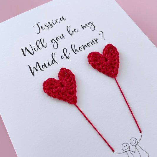 A white card with a line drawn couple printed on it - the couple are each holding a red crochet heart balloon. Will you be my Maid of honour is printed at the top of the card with the option of adding a name, making it the perfect keepsake. 