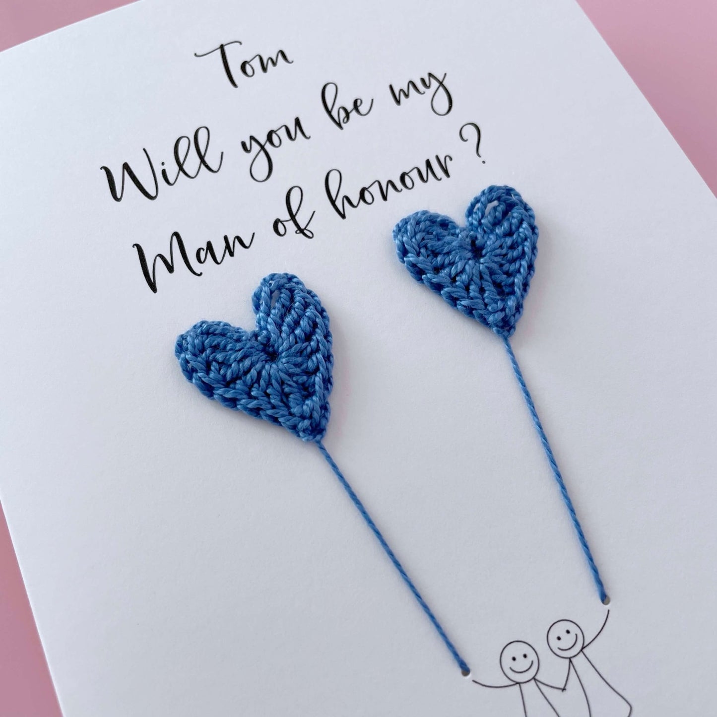 A white card with a line drawn couple printed on it - the couple are each holding a blue crochet heart balloon. Will you be my Man of honour is printed at the top of the card with the option of adding a name, making it the perfect keepsake. 