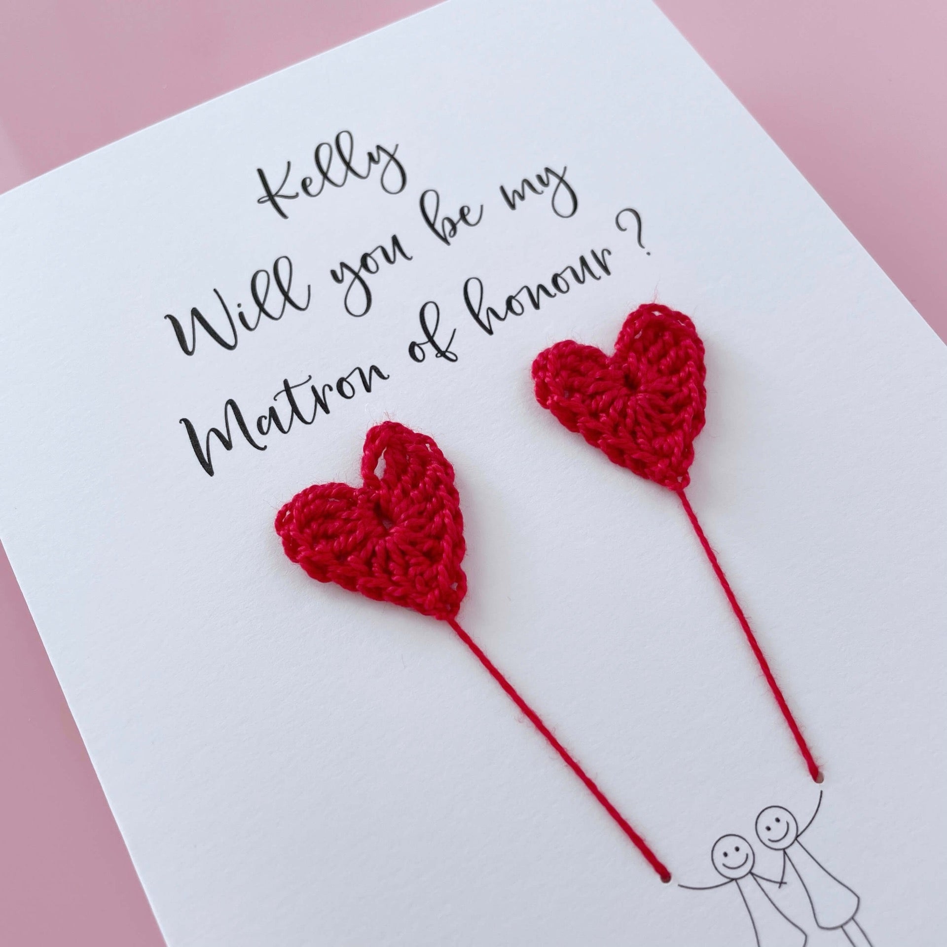 A white card with a line drawn couple printed on it - the couple are each holding a red crochet heart balloon. Will you be my Matron of honour is printed at the top of the card with the option of adding a name, making it the perfect keepsake. 