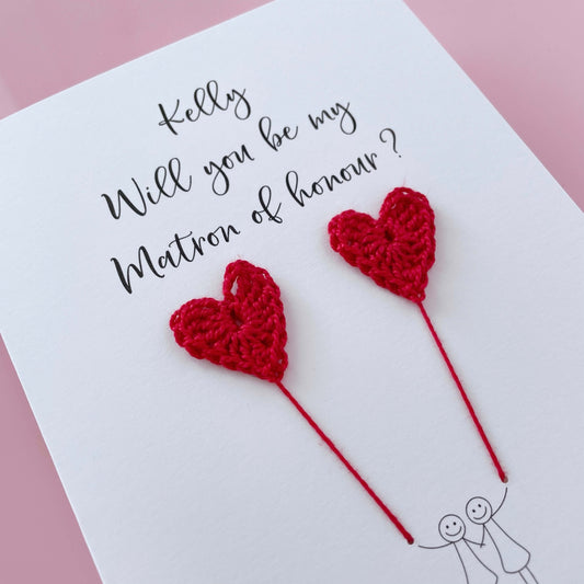 A white card with a line drawn couple printed on it - the couple are each holding a red crochet heart balloon. Will you be my Matron of honour is printed at the top of the card with the option of adding a name, making it the perfect keepsake. 
