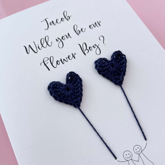 A white card with a line drawn couple printed on it - the couple are each holding a Navy blue crochet heart balloon. Will you be our Flower boy is printed at the top of the card with the option of adding a name, making it the perfect keepsake. 