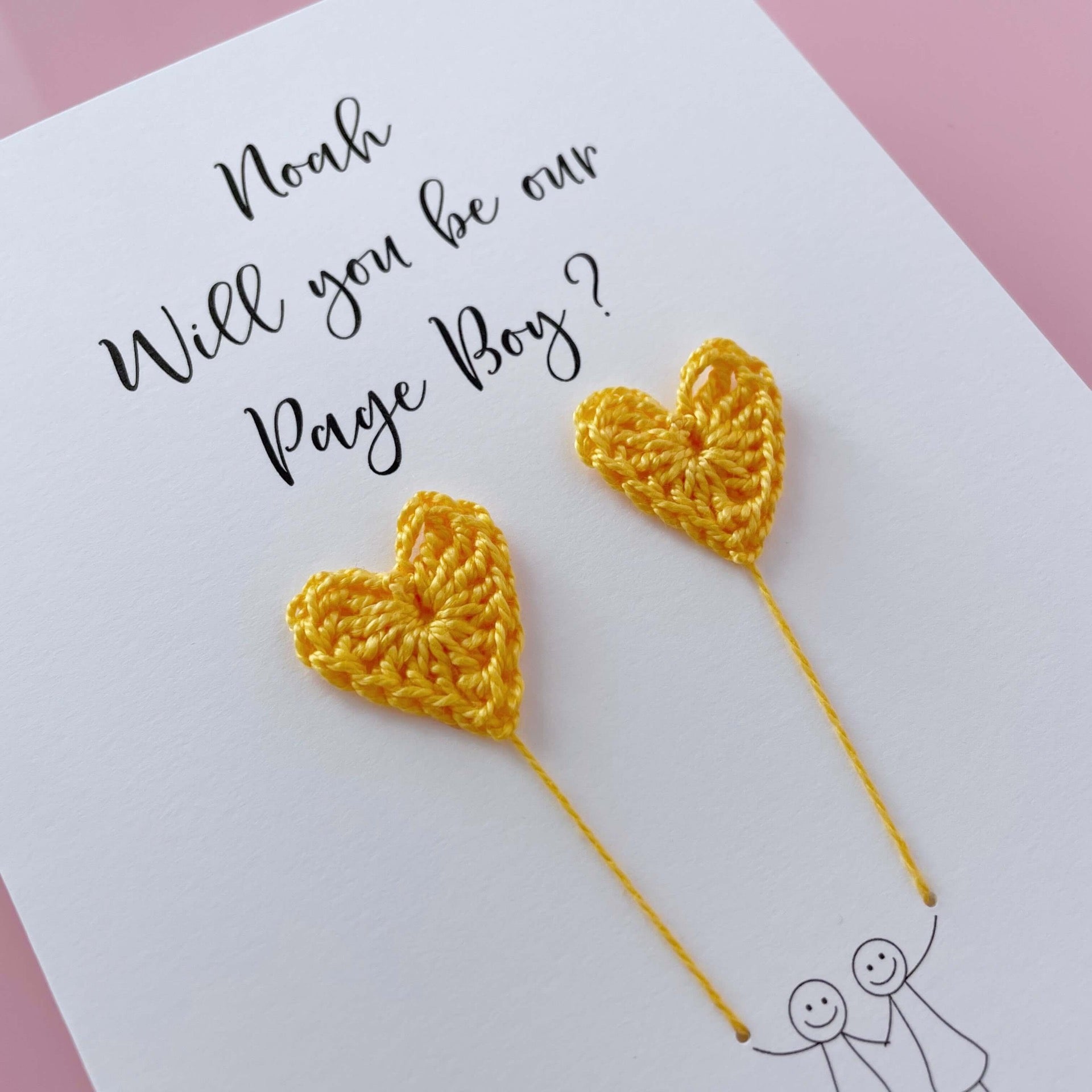 A white card with a line drawn couple printed on it - the couple are each holding a yellow crochet heart balloon. Will you be our page boy is printed at the top of the card with the option of adding a name, making it the perfect keepsake. 