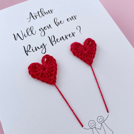 A white card with a line drawn couple printed on it - the couple are each holding a red crochet heart balloon. Will you be our Ring bearer is printed at the top of the card with the option of adding a name, making it the perfect keepsake. 