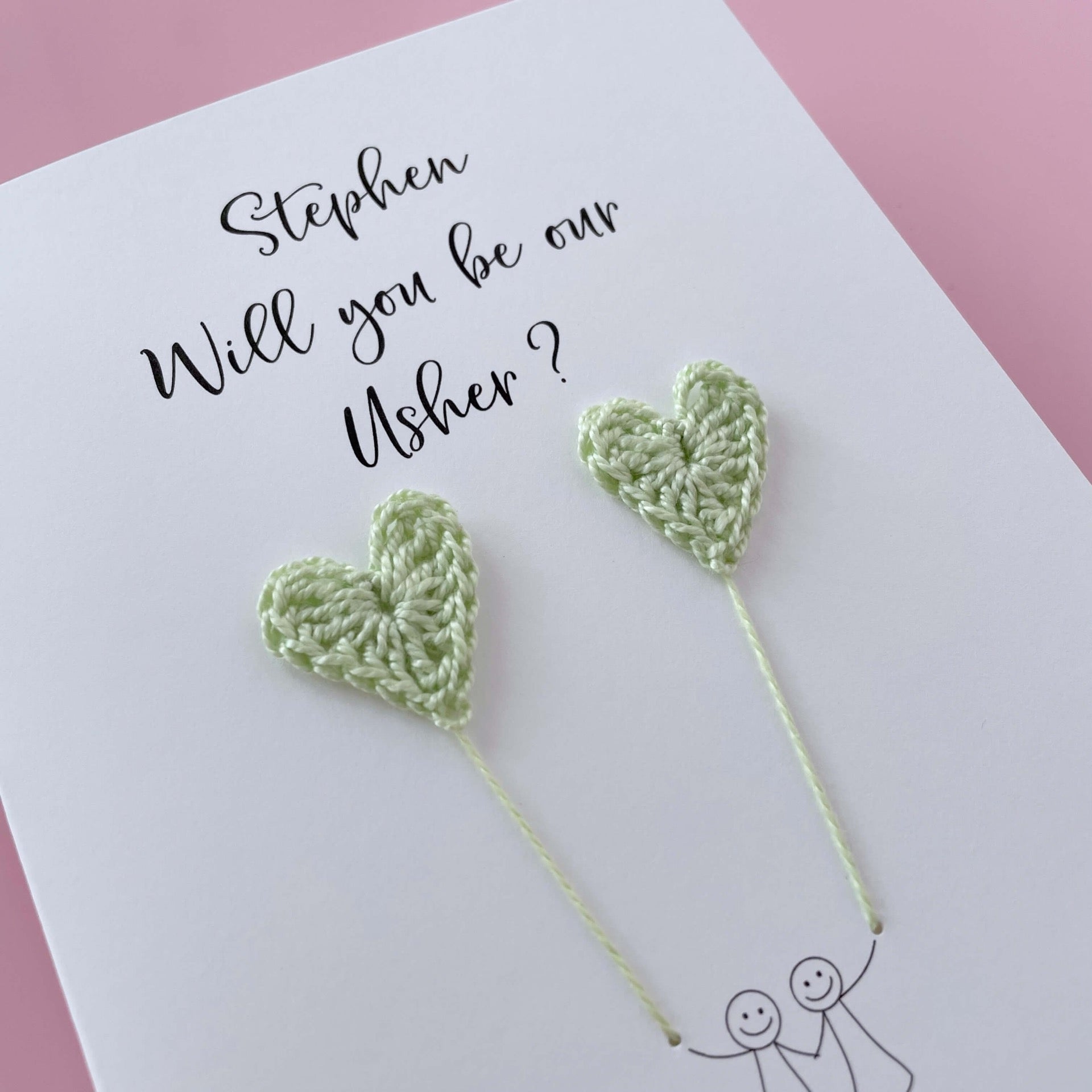 A white card with a line drawn couple printed on it - the couple are each holding a pale green crochet heart balloon. Will you be our Usher is printed at the top of the card with the option of adding a name, making it the perfect keepsake. 