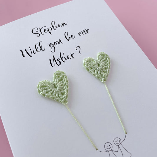 A white card with a line drawn couple printed on it - the couple are each holding a pale green crochet heart balloon. Will you be our Usher is printed at the top of the card with the option of adding a name, making it the perfect keepsake. 