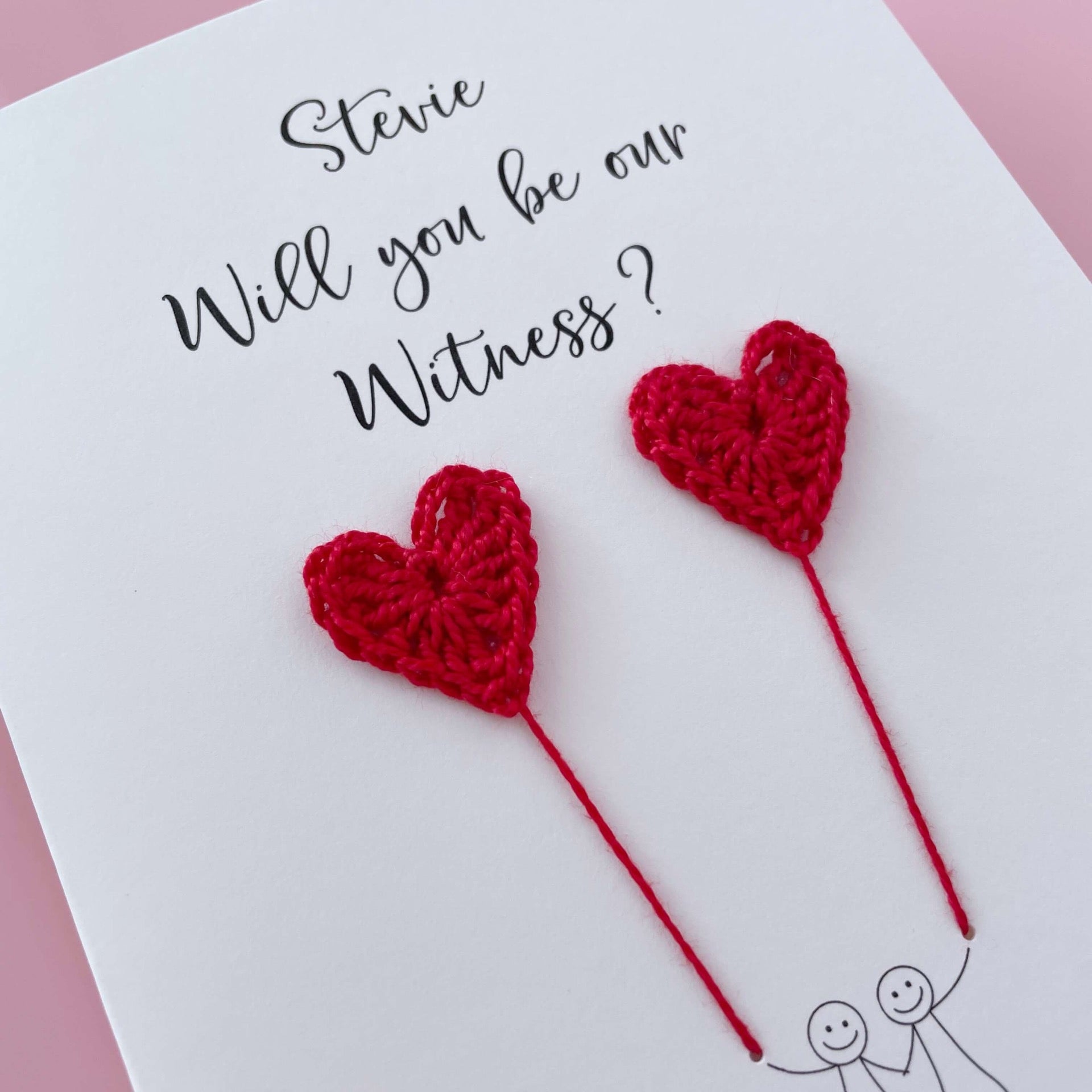 A white card with a line drawn couple printed on it - the couple are each holding a Red crochet heart balloon. Will you be our witness is printed at the top of the card with the option of adding a name, making it the perfect keepsake. 