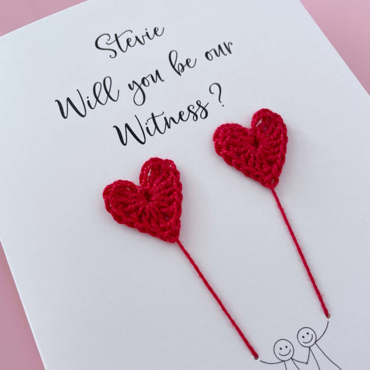 A white card with a line drawn couple printed on it - the couple are each holding a Red crochet heart balloon. Will you be our witness is printed at the top of the card with the option of adding a name, making it the perfect keepsake. 